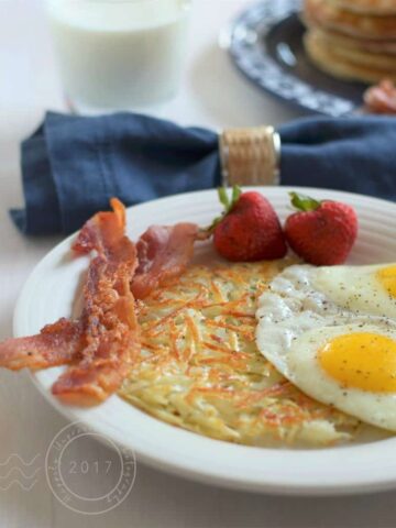 White plate with crispy golden shredded hashbrowns, 2 eggs sunny side up, 2 slices bacon, 2 strawberries with blue napkin and glass of milk