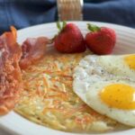 White plate with crispy golden shredded hashbrowns, 2 eggs sunny side up, 2 slices bacon, 2 strawberries with blue napkin and glass of milk