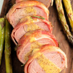 Bacon Wrapped Pork Tenderloin sliced on a cutting board drizzled in a honey dijon sauce