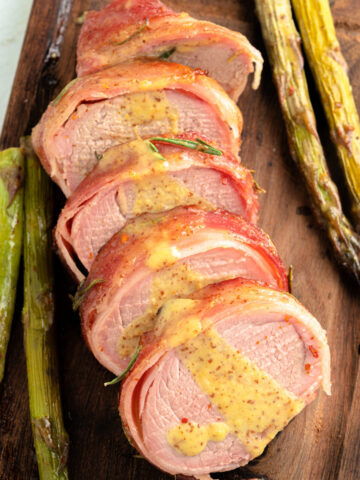 Bacon Wrapped Pork Tenderloin sliced on a cutting board drizzled in a honey dijon sauce