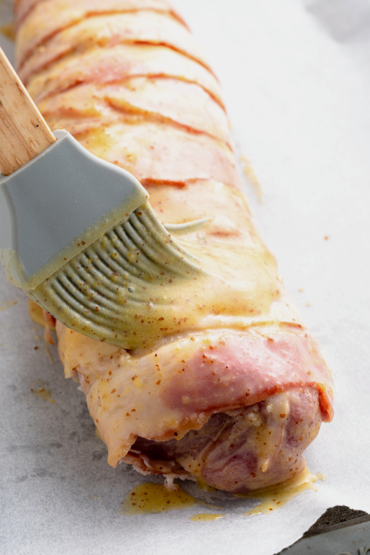 A pork tenderloin wrapped in bacon being brushed in a Honey Dijon glaze before cooking